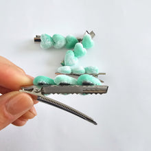 A silver metal 5.7cm alligator clip decorated with velvety, pastel mint green hearts, held by a person's hand against a plain white background.