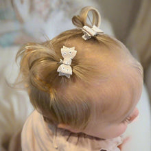 Young blonde preschool aged child with hair styled in tiny pigtails with decorative bow hair clips in beige with a white and black floral print, wearing a light-colored top.