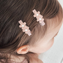 A brunette toddler child's head with two dusky pink ribbon wrapped alligator style hair clips adorned with lace flowers.
