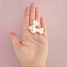 Two pale beige faux-leatger hair bows on 2.5cm clips with held in a hand against a pink background