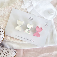 Three bow-shaped hair clips by Ruby & Bear are displayed on a branded white card, styled on soft white lace fabric with decorative shoes partially visible at the edges.