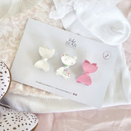 Three bow-shaped hair clips by Ruby & Bear are displayed on a branded white card, styled on soft white lace fabric with decorative shoes partially visible at the edges.