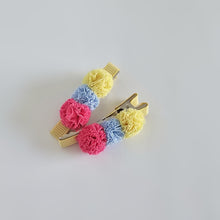 Two silver coloured alligator hair clips wrapped in buttery yellow grosgrain ribbon, embellished with soft, nylon mesh pom-poms in yellow, light blue, and bright pink, arranged on a light gray background.