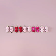 A selection of heart-shaped hair clips in various colors, including red, pastel pink, and hot pink, laid out on a pink background.
