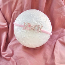 A light pink fabric headband with a decorative bow featuring a pink and white floral pattern, displayed on a textured white circular prop over a draped pink satin fabric background.