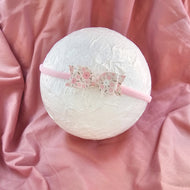 A light pink fabric headband with a decorative bow featuring a pink and white floral pattern, displayed on a textured white circular prop over a draped pink satin fabric background.
