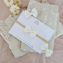 Beige baby clothes in a pile with soft white bow headband and hairclips on a pink background