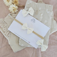 Beige baby clothes in a pile with soft white bow headband and hairclips on a pink background