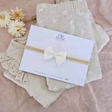 Beige baby clothes laid in a pile underneath a soft white, faux suede bow sewn onto a stretchy headband, made by Ruby and Bear NZ, on a pale pink background