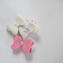 Three faux leather hair bows on a light gray background: one solid soft white bow, one soft white bow with a pink and green floral print, and one solid pink bow.
