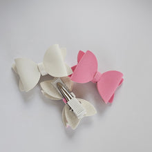 Three small bow hair clips made in New Zealand by Ruby & Bear on a light gray background: one soft white, one pink, and one white with a small floral print accent visible.